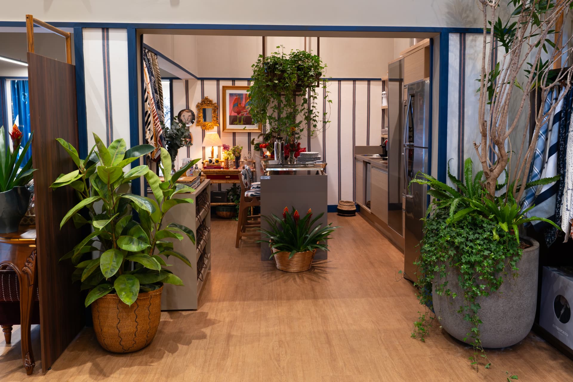 Entrada do ambiente Comporta com paredes listradas em azul, cozinha ao fundo e grandes vasos de plantas