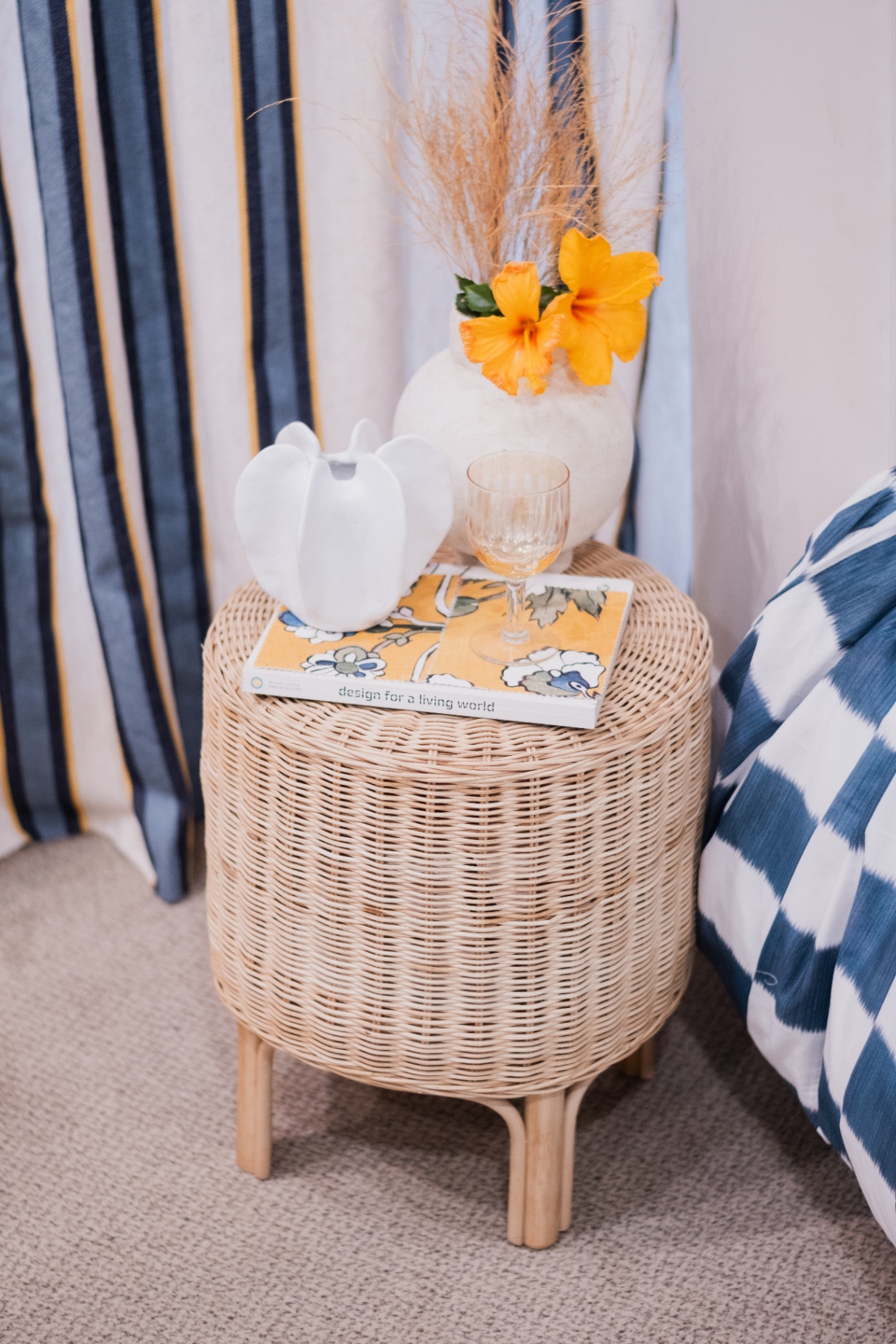 Detalhe de banqueta de vime com vaso branco, flor de hibisco amarelo e livro sobre design, com cortinas listradas ao fundo