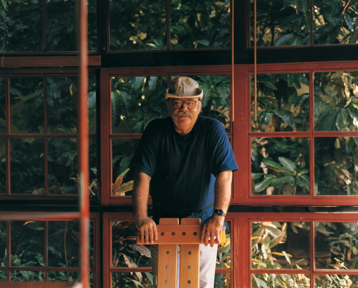 Retrato de Sergio Rodrigues, designer brasileiro, encostado em cadeira de madeira diante de janelas de esquadria vermelha e vegetação