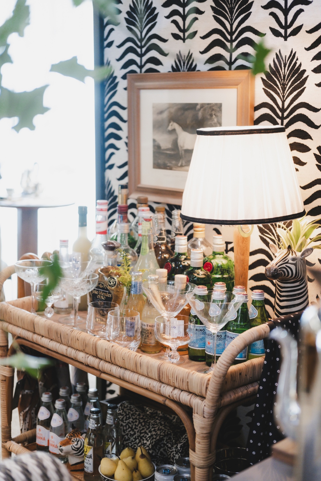 Bar cart de vime com garrafas, taças de cristal e abajur ao lado de papel de parede zebra Entreposto