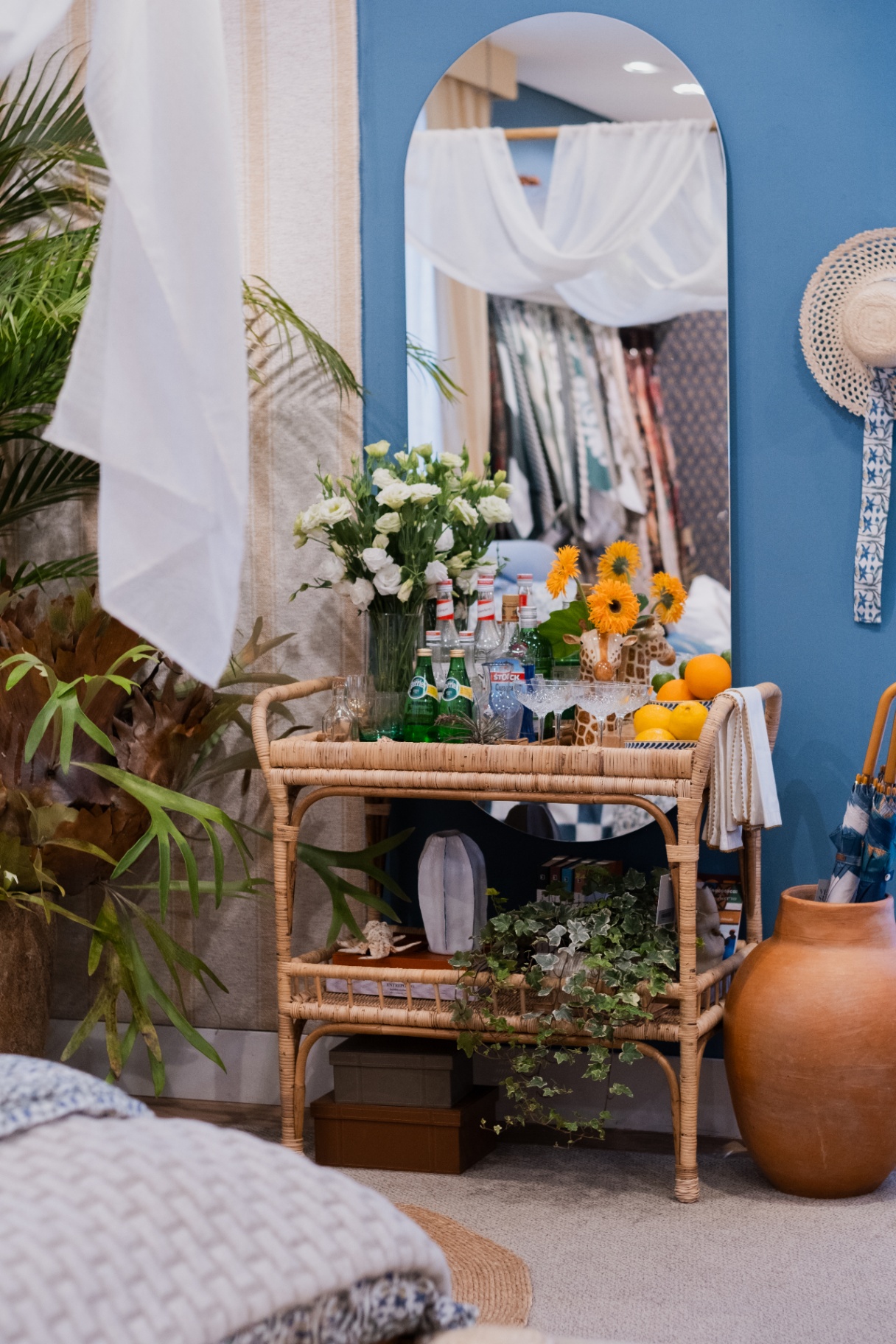 Bar cart de vime com flores, taças e frutas diante de parede azul e cortina listrada da coleção Zanzibar