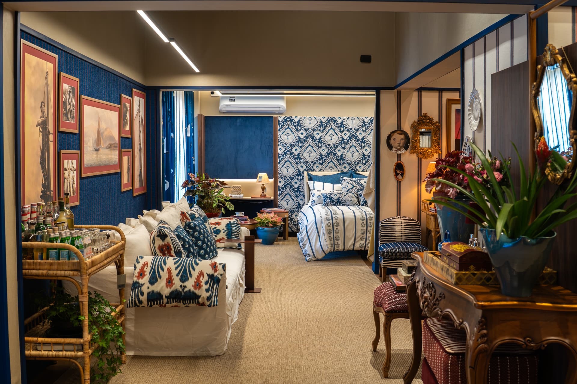 Vista do ambiente Comporta com sofá claro, almofadas de estampa ikat azul e vermelha, galeria de quadros e quarto ao fundo