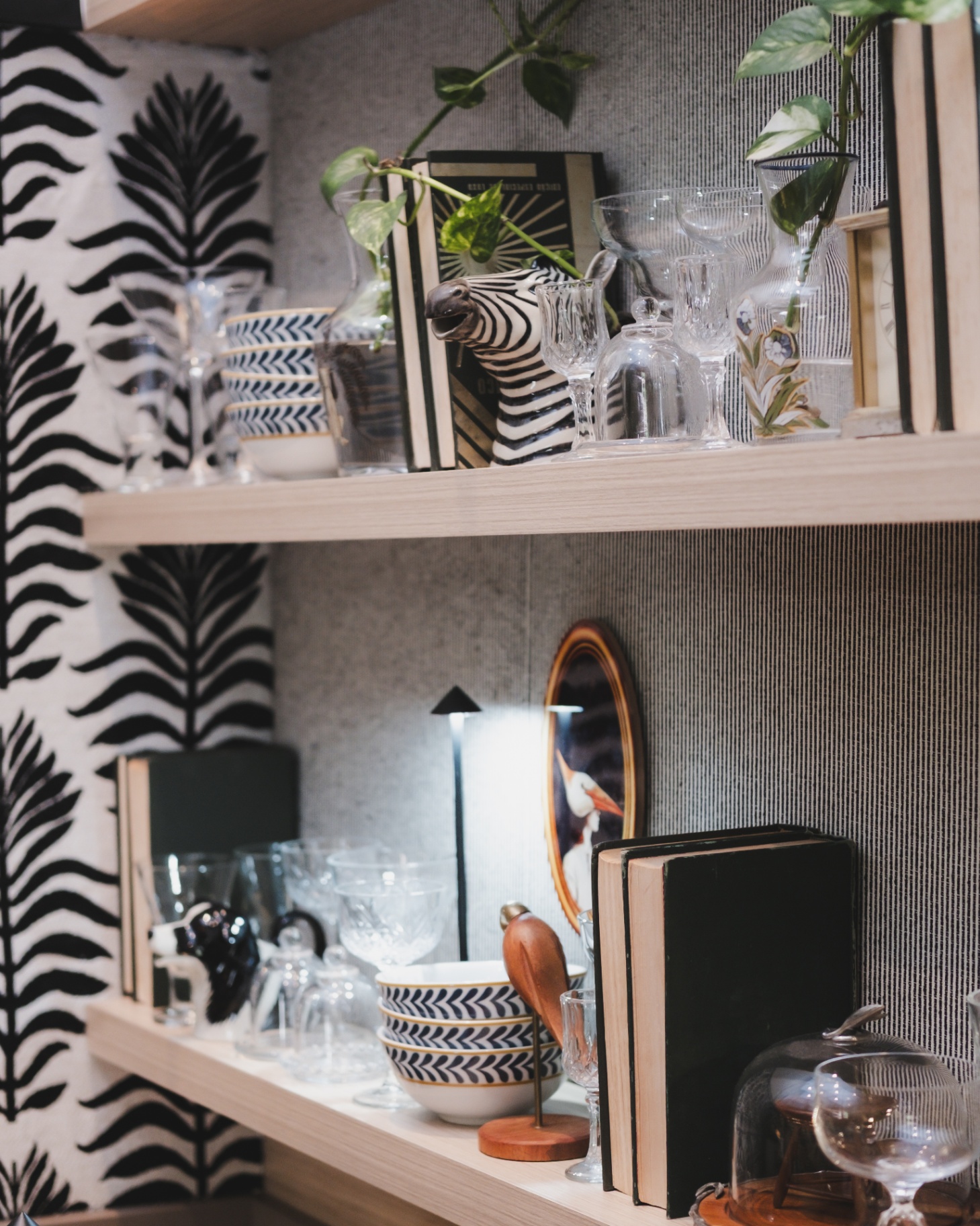 Estante com louças cheurom, cristais e escultura de zebra sobre fundo de papel de parede preto e branco