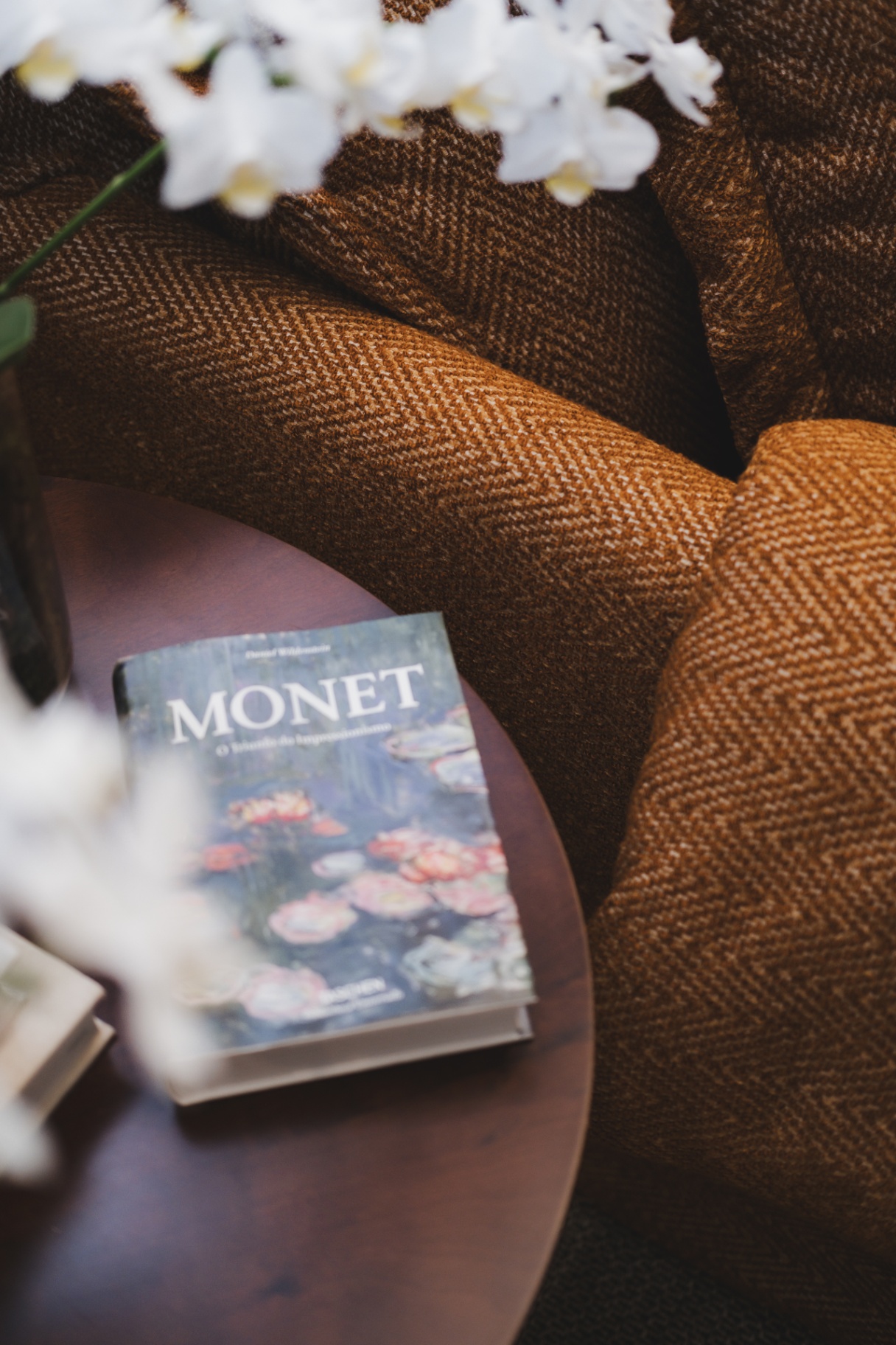 Livro Monet sobre mesa lateral, entre estofado texturizado e arranjo de orquídeas brancas