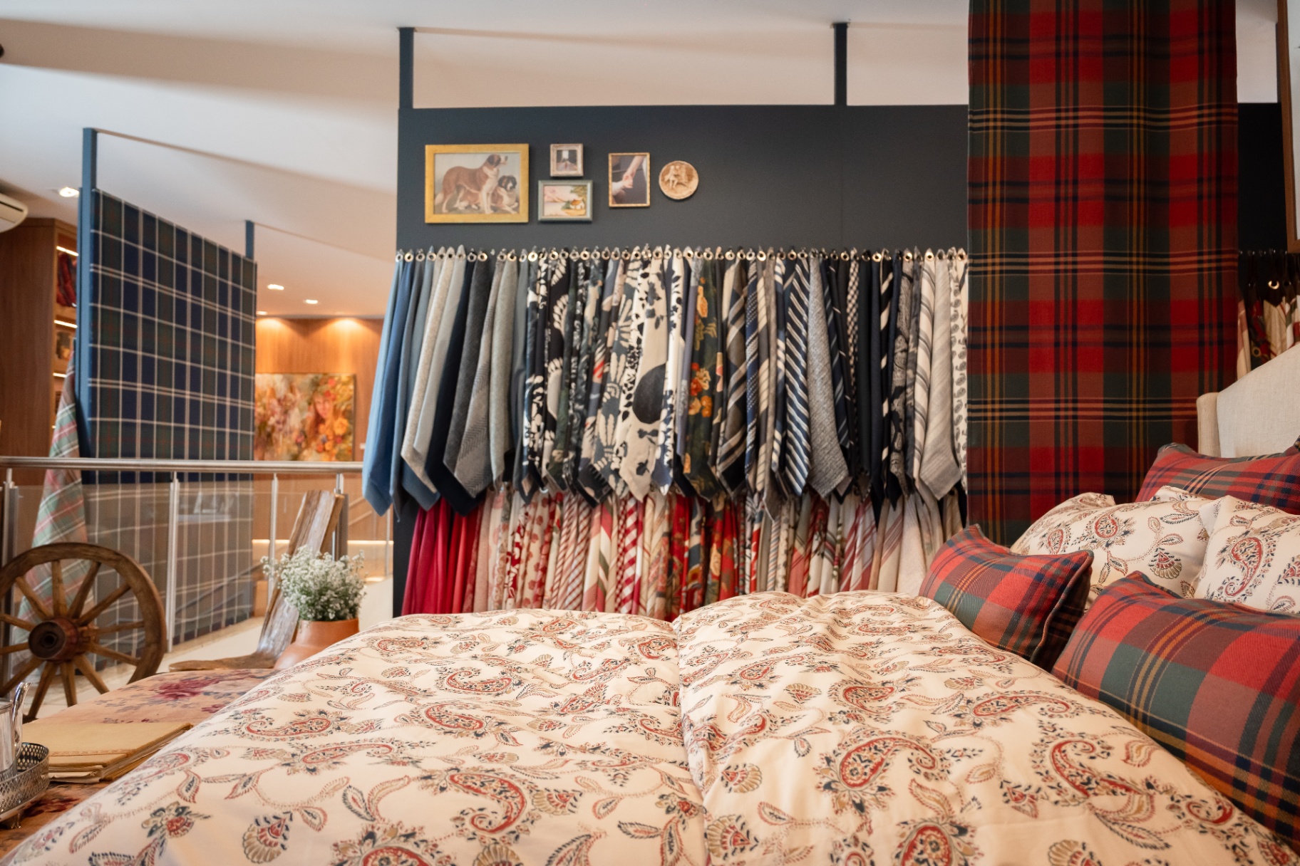 Cama com roupa de cama paisley e almofadas xadrez, entre araras de tecidos Entreposto