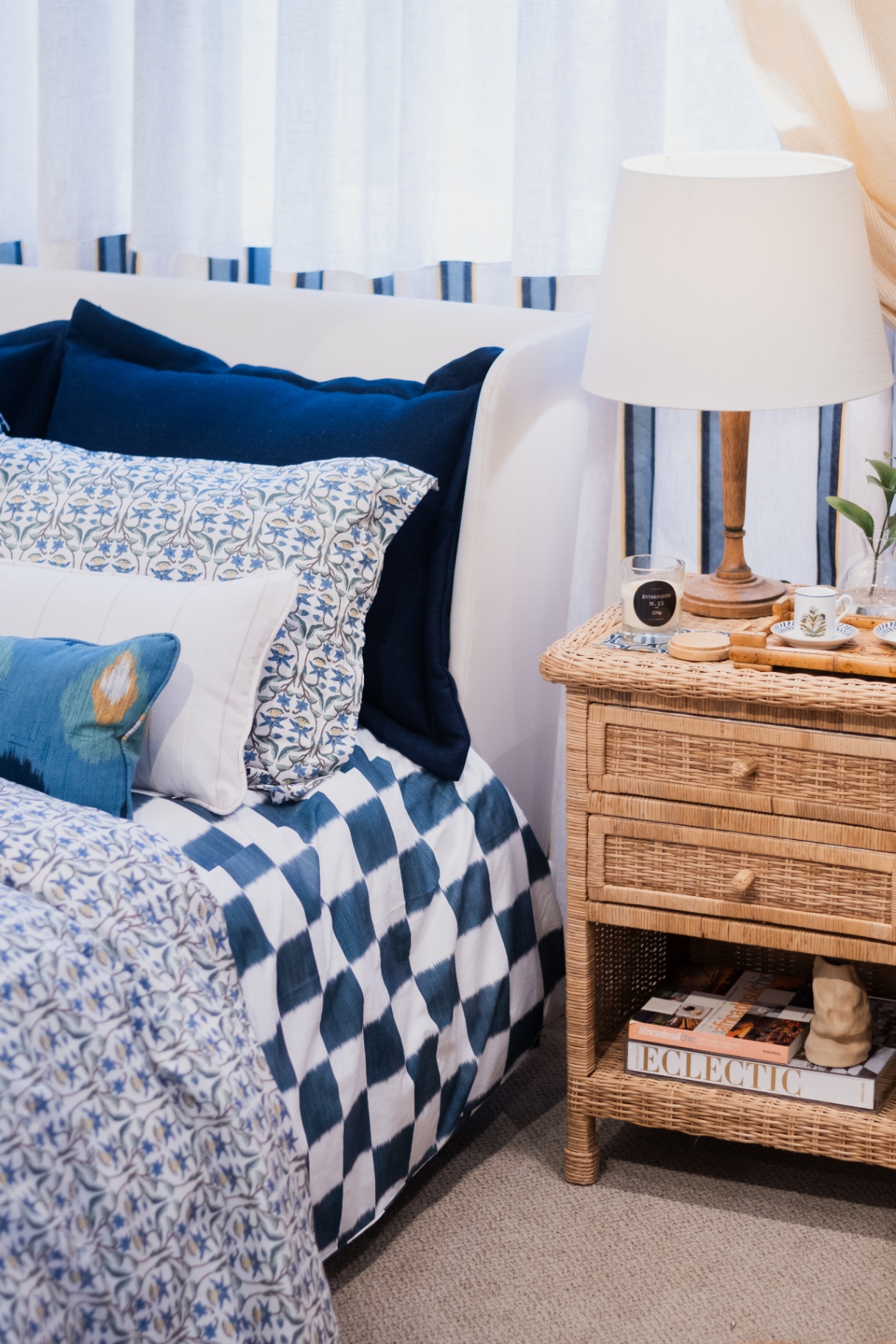 Cabeceira branca com roupa de cama azul estampada, almofadas de linho e abajur de cúpula sobre criado de vime